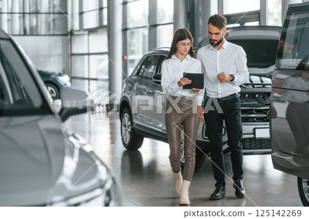 Front view. Man with woman in white clothes are in the car dealership together 125142269