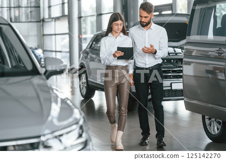 Front view. Man with woman in white clothes are in the car dealership together 125142270