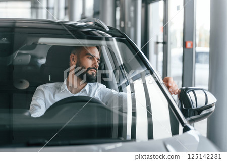 Front view. Sitting inside of automobile. Young man in white clothes is in the car dealership 125142281