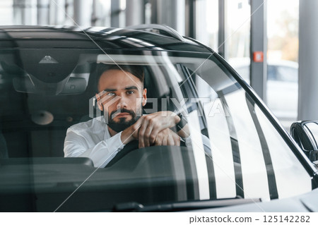 Front view. Sitting inside of automobile. Young man in white clothes is in the car dealership Front view. Sitting inside of automobile. Young man in white clothes is in the car dealership 125142282