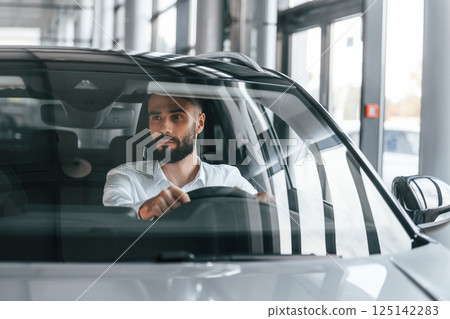 Front view. Sitting inside of automobile. Young man in white clothes is in the car dealership 125142283