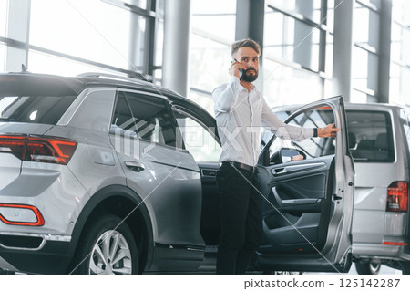 Talking by phone. Young man in white clothes is in the car dealership Talking by phone. Young man in white clothes is in the car dealership 125142287