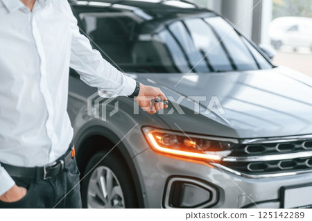Using the keys remote controller. Young man in white clothes is in the car dealership Using the keys remote controller. Young man in white clothes is in the car dealership 125142289