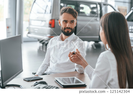 Owner of new automobile is receiving keys. Man with woman in white clothes are in the car dealership together 125142304