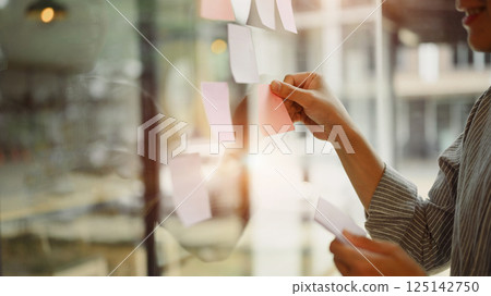 Businessman placing colorful paper notes on transparent board during team strategy session Businessman placing colorful paper notes on transparent board during team strategy session 125142750