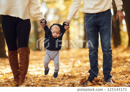 Holding toddler by the hands. Two parents are having a walk with their little son in the autumn park Holding toddler by the hands. Two parents are having a walk with their little son in the autumn park 125143171