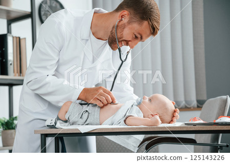 Close up view. Using stethoscope. Doctor in white coat is taking care of little toddler indoors 125143226