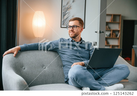 Sitting and looking at the side. Male freelance worker is using laptop at home. Cozy apartment Sitting and looking at the side. Male freelance worker is using laptop at home. Cozy apartment 125143262