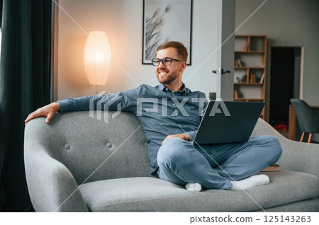 Sitting and looking at the side. Male freelance worker is using laptop at home. Cozy apartment Sitting and looking at the side. Male freelance worker is using laptop at home. Cozy apartment 125143263