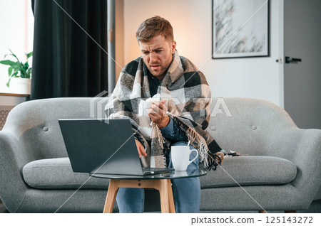 Sitting and sneezes. Sick man is on the sofa at home by the laptop. Conception of illness Sitting and sneezes. Sick man is on the sofa at home by the laptop. Conception of illness 125143272