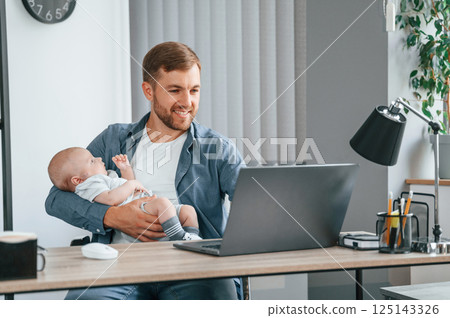 Working by using laptop. Holding baby. Father with toddler is indoors in the office 125143326