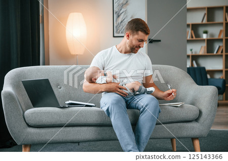 Sitting on the sofa and using pen. Father with toddler is at home, taking care of his son Sitting on the sofa and using pen. Father with toddler is at home, taking care of his son 125143366