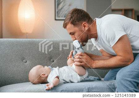 Baby is laying down on the sofa. Father with toddler is at home, taking care of his son Baby is laying down on the sofa. Father with toddler is at home, taking care of his son 125143389