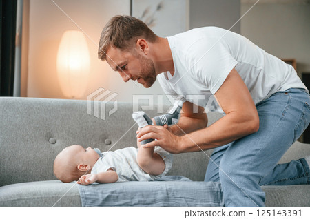 Baby is laying down on the sofa. Father with toddler is at home, taking care of his son 125143391