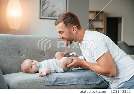 Baby is laying down on the sofa. Father with toddler is at home, taking care of his son 125143392