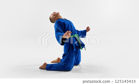 Martial artist celebrates victory on knees, fists clenched, head raised in triumph, expressing emotional release, pride, and power in competitive judo success. 125143433
