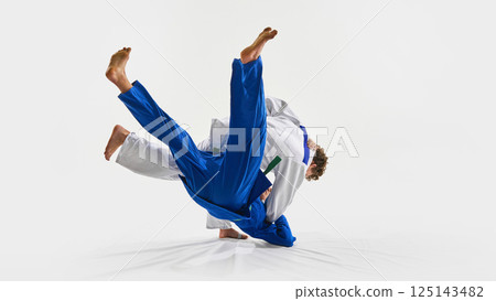 Powerful judo throw in motion as blue gi athlete flips mid-air, while opponent maintains grip, symbolizing energy against white background. Powerful judo throw in motion as blue gi athlete flips mid-air, while opponent maintains grip, symbolizing energy against white background. 125143482