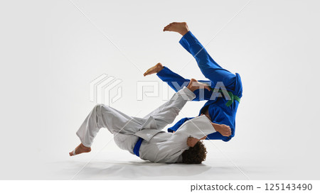 Ground-level flip from judoka in white as fighter in blue is thrown backwards, legs high in air, showing timing, discipline against white background. 125143490