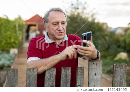 Mature man standing relaxed near fence talking on phone in backyard smiling Mature man standing relaxed near fence talking on phone in backyard smiling 125143495