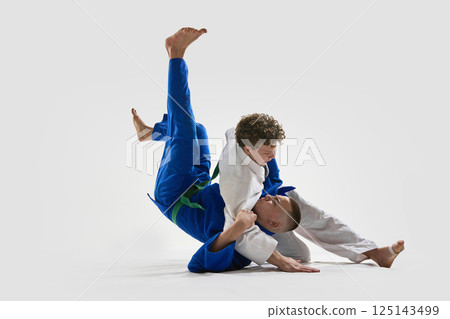 Young judoka in white holds opponent in blue during intense ground control, demonstrating control, and strength against white background. 125143499