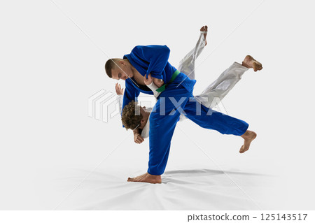 Judoka in white executes forward throw, lifting opponent over shoulder in full motion, reflecting strength, precision against white studio background. Judoka in white executes forward throw, lifting opponent over shoulder in full motion, reflecting strength, precision against white studio background. 125143517