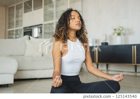 Yoga mindfulness meditation. Young healthy African girl practicing yoga at home. Woman sitting in lotus pose on yoga mat meditating relaxing indoor. Girl doing breathing practice. Yoga at home 125143540