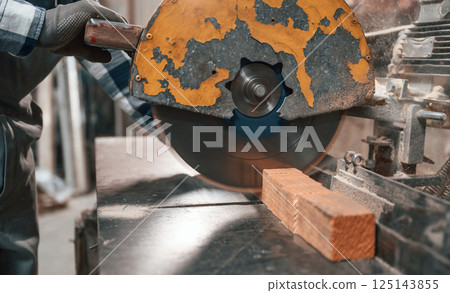Cutting the wood by saw. Factory male worker in uniform is indoors 125143855