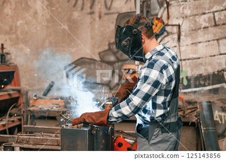 In protective mask. Welding the iron. Factory male worker in uniform is indoors 125143856