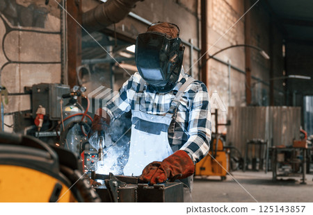 In protective mask. Welding the iron. Factory male worker in uniform is indoors 125143857