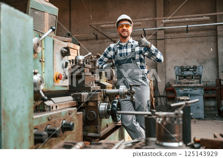 Leaning on the factory machine and showing thumb up. Worker in uniform is indoors 125143929