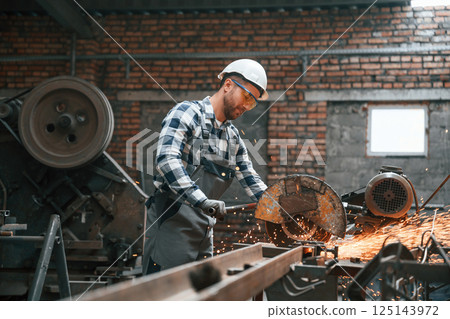 Factory male worker in uniform and hard hat is indoors, cutting the metal 125143972