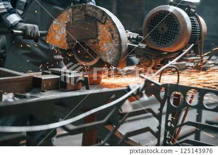 Many of the sparks. Cutting metal. Factory male worker in uniform is indoors Many of the sparks. Cutting metal. Factory male worker in uniform is indoors 125143976