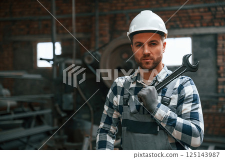 Standing in grey uniform and in white hard hat. Factory male worker is indoors 125143987