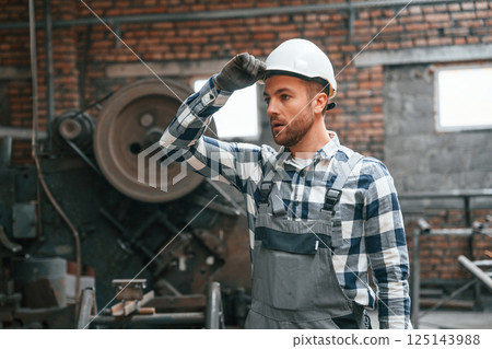 Standing in grey uniform and in white hard hat. Factory male worker is indoors 125143988