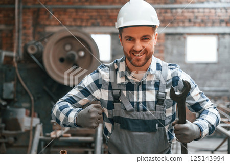 Standing and showing wrench. Factory male worker in uniform is indoors Standing and showing wrench. Factory male worker in uniform is indoors 125143994