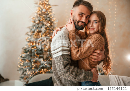 Standing against the fir with garlands on it. Lovely young couple are celebrating New Year at home Standing against the fir with garlands on it. Lovely young couple are celebrating New Year at home 125144121