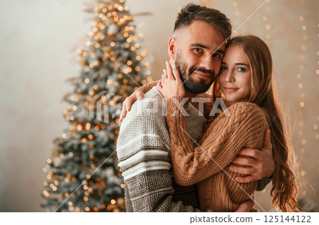 Standing against the fir with garlands on it. Lovely young couple are celebrating New Year at home Standing against the fir with garlands on it. Lovely young couple are celebrating New Year at home 125144122