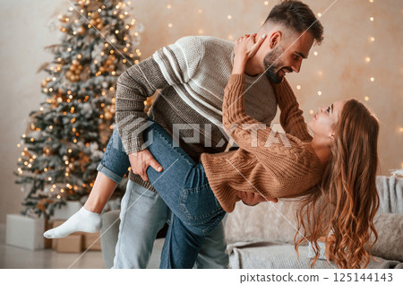 Man is holding leg of the woman while standing. Lovely young couple are celebrating New Year at home 125144143