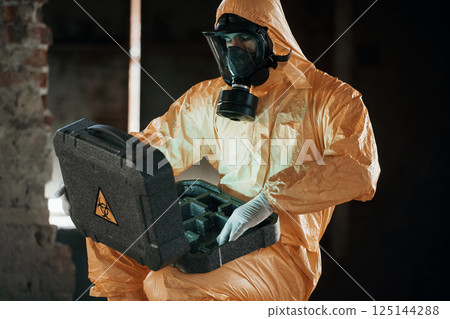 Case with blue glowing light. Hazard materials. Man dressed in chemical protection suit in the ruins of the post apocalyptic building 125144288