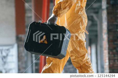 The case with biohazard sign on it. Man dressed in chemical protection suit in the ruins of the post apocalyptic building 125144294