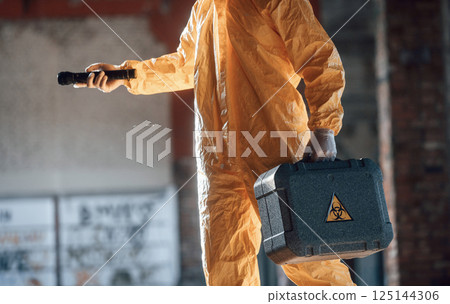 Holding case and flashlight. Man dressed in chemical protection suit in the ruins of the post apocalyptic building 125144306