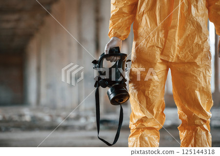 Holding gas mask. Man dressed in chemical protection suit in the ruins of the post apocalyptic building 125144318