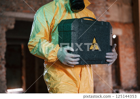Case with biohazard sign in hands. Man dressed in chemical protection suit in the ruins of the post apocalyptic building Case with biohazard sign in hands. Man dressed in chemical protection suit in the ruins of the post apocalyptic building 125144353