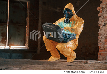 Case with blue glowing light. Hazard materials. Man dressed in chemical protection suit in the ruins of the post apocalyptic building Case with blue glowing light. Hazard materials. Man dressed in chemical protection suit in the ruins of the post apocalyptic building 125144361