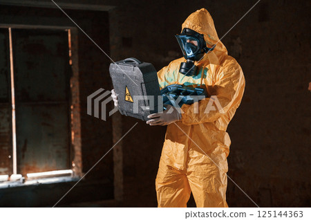 Blue light is going from hard case that is with hazard sign. Man dressed in chemical protection suit in the ruins of the post apocalyptic building 125144363