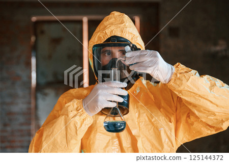 Holding a test tube and examining radioactive liquid. Man dressed in chemical protection suit in the ruins of the post apocalyptic building Holding a test tube and examining radioactive liquid. Man dressed in chemical protection suit in the ruins of the post apocalyptic building 125144372