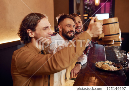 Raising up the mugs. Three friends are in the bar drinking beer and having a good time Raising up the mugs. Three friends are in the bar drinking beer and having a good time 125144697