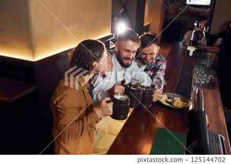 Sitting by the table. Three friends are in the bar drinking beer and having a good time Sitting by the table. Three friends are in the bar drinking beer and having a good time 125144702