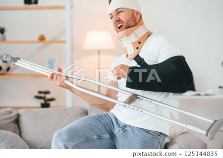 Standing, holding crutch and pretending that he's playing guitar. Having fun. Man is at home indoors. Having injuries 125144835