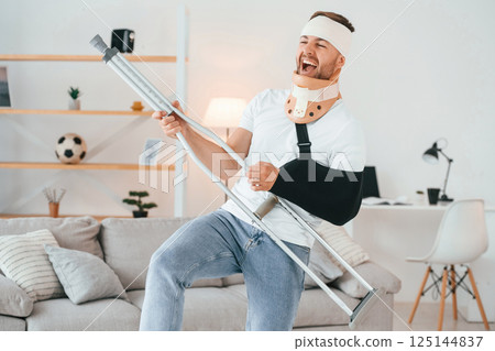 Standing, holding crutch and pretending that he's playing guitar. Having fun. Man is at home indoors. Having injuries 125144837
